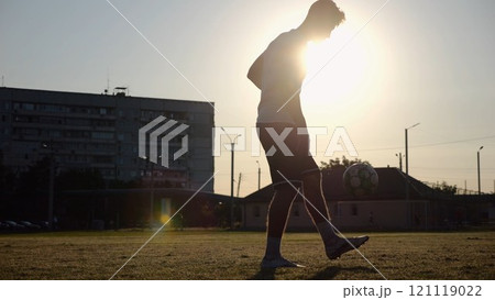 Professional footballer juggling soccer ball on stadium at sunset. Young man kicking ball at green field. Sportsman practicing tricks at meadow with sunlight at background. Freestyle football concept 121119022