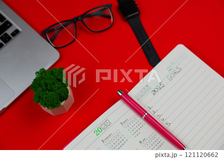 A close-up on a red background with a slope lies a pink diary notebook in which it is written: Goals for 2025, inside it is a calendar for 2025 and there is a pink pen, an open laptop, part of the A close-up on a red background with a slope lies a pink diary notebook in which it is written: Goals for 2025, inside it is a calendar for 2025 and there is a pink pen, an open laptop, part of the 121120662