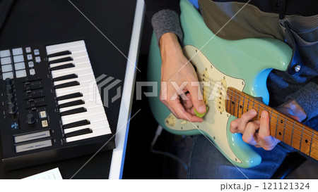 Male musician skillfully strums a mint electric guitar while a MIDI keyboard controller sits nearby during a songwriting session in his home studio 121121324