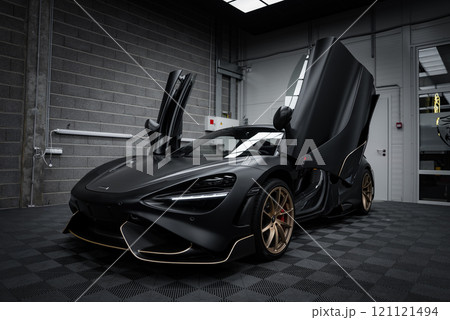 A matte black McLaren 720S with open dihedral doors is set in a modern garage. The car features gold accented wheels and a checkered floor backdrop. A matte black McLaren 720S with open dihedral doors is set in a modern garage. The car features gold accented wheels and a checkered floor backdrop. 121121494