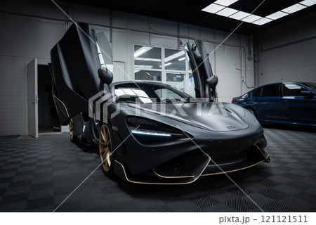 The McLaren 720S is parked indoors on a checkered floor, showcasing its matte black finish, gold accents, and distinctive dihedral doors under bright lighting. The McLaren 720S is parked indoors on a checkered floor, showcasing its matte black finish, gold accents, and distinctive dihedral doors under bright lighting. 121121511