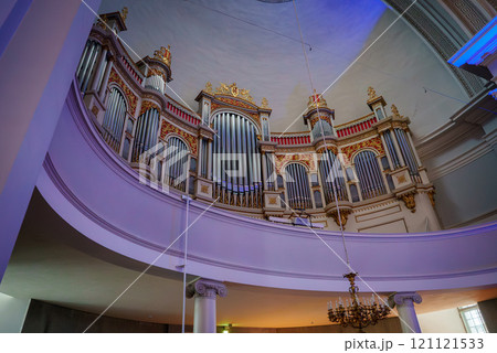 A grand pipe organ with gold and red accents is set against white columns and a curved balcony in a Helsinki church. A chandelier hangs from the domed ceiling. 121121533
