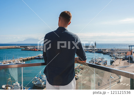 A man stands on a balcony overlooking a marina filled with boats, with a lighthouse at the pier's end. Distant mountains and ocean under a blue sky. 121121536