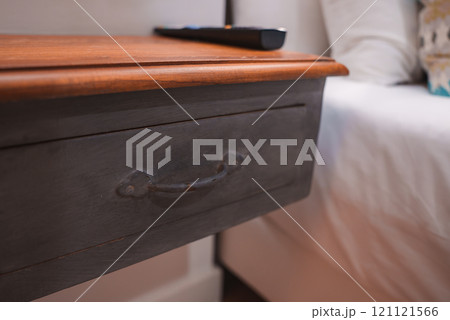 A wooden bedside table with a dark finish and metal handle drawer. A remote control rests on its lighter wood surface. White linens and a pillow are visible. 121121566