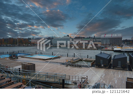 View of Helsinki's waterfront featuring a wooden deck, small structures, and seating areas. The skyline includes a mix of modern and traditional architecture. 121121642