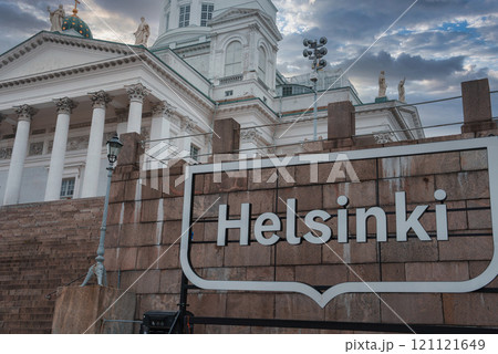 Helsinki Cathedral's neoclassical architecture stands tall with white columns and statues, contrasted by a modern sign in the foreground under a dramatic sky. 121121649