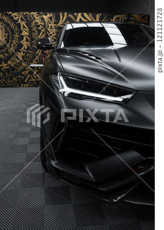 The image captures the front end of a Lamborghini Urus Performante with a matte black finish, set against a patterned wall indoors, highlighting its design. The image captures the front end of a Lamborghini Urus Performante with a matte black finish, set against a patterned wall indoors, highlighting its design. 121121728