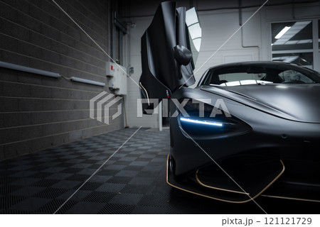 The McLaren 720S is displayed indoors on a checkered floor, featuring its aerodynamic design and LED headlights. A brick wall and piping are visible. 121121729