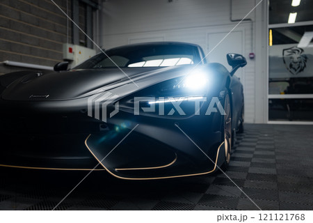 The McLaren 720S is parked indoors on a checkered floor, highlighting its aerodynamic design, LED headlights, and matte black finish in a modern facility. 121121768