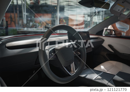 The image captures a modern car interior with a sleek steering wheel and large touchscreen. Through the windows, blurred glass structures and greenery suggest an urban setting. 121121770