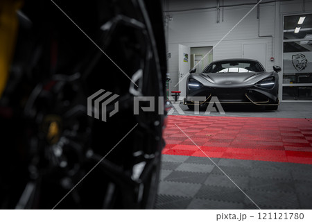 A McLaren 720S is parked in a modern garage with a red and black checkered floor. The car's aerodynamic design and sharp headlights are highlighted, with a blurred Lamborghini Urus wheel in the 121121780