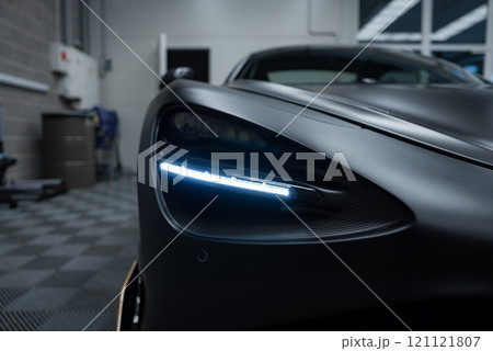 The image captures the sleek design of a McLaren 720S headlight with a distinctive LED strip. The matte black finish is set against a checkered floor. The image captures the sleek design of a McLaren 720S headlight with a distinctive LED strip. The matte black finish is set against a checkered floor. 121121807