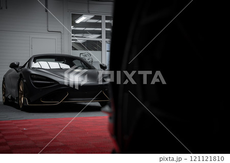 A matte black McLaren 720S is parked indoors on a red carpeted floor, with white walls and large windows. The car's aerodynamic design is highlighted. A matte black McLaren 720S is parked indoors on a red carpeted floor, with white walls and large windows. The car's aerodynamic design is highlighted. 121121810