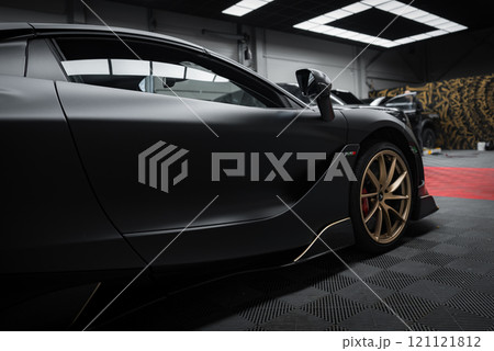 The image features a McLaren 720S indoors, highlighting its sleek design, matte black finish, and gold alloy wheels. A Lamborghini Urus Performante is partially visible in the background. 121121812