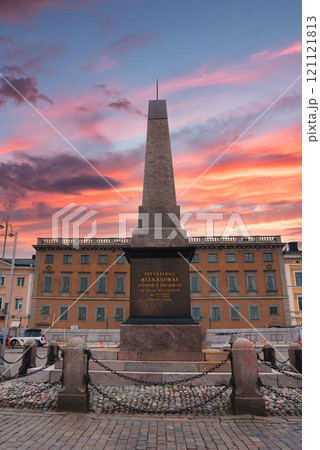 The Keisarinnankivi obelisk stands in Helsinki's Market Square, framed by a vibrant sunset sky. A European style building adds cultural context. The Keisarinnankivi obelisk stands in Helsinki's Market Square, framed by a vibrant sunset sky. A European style building adds cultural context. 121121813