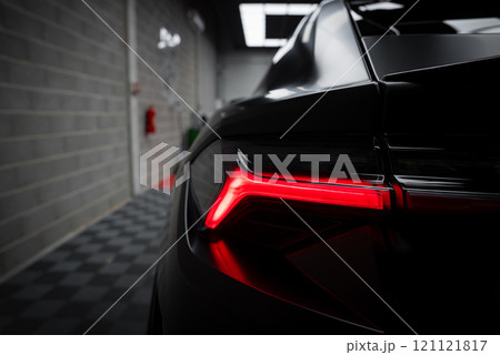 The image captures the angular design of a supercar's taillight, contrasting red light with a dark glossy body. Tiled floor and brick walls are visible. 121121817