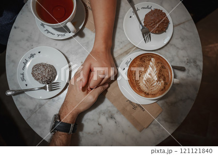 A marble table in a Helsinki cafe features cappuccino with latte art, tea, and desserts. Two hands clasp in the center, creating an intimate scene. 121121840