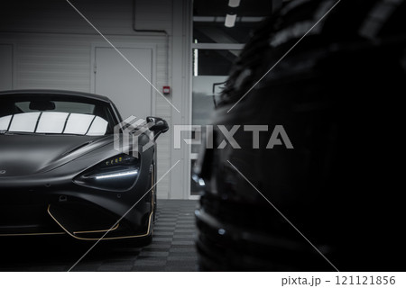 The McLaren 720S is displayed indoors, highlighting its matte black finish, gold accents, and LED headlights. A blurred vehicle adds depth to the scene. 121121856