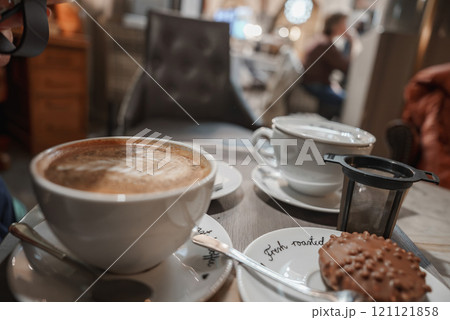 Close up of a cafe table in Helsinki with two coffee cups and a dessert. The scene includes plates labeled 'fresh roasted' and a blurred interior with patrons. 121121858