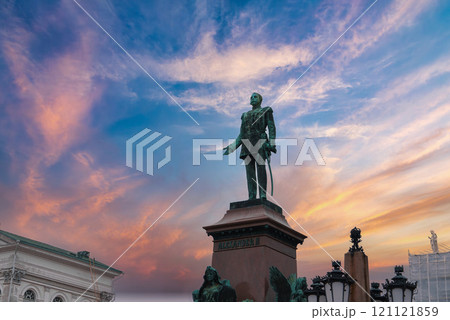 The statue of Alexander II stands in Senate Square, Helsinki, against a vibrant sunset sky. Helsinki Cathedral is partially visible on the left. 121121859