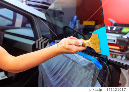 Hand worker carefully applies squeegee to install window film on vehicle side window in workshop environment. 121121917