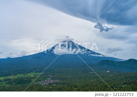 Panorama of Mount Agung and rice fields on the island of Bali. View of the mountain against a background of palm trees and a cornfield. Panorama of Agung volcano covered with clouds on a rainy day. Panorama of Mount Agung and rice fields on the island of Bali. View of the mountain against a background of palm trees and a cornfield. Panorama of Agung volcano covered with clouds on a rainy day. 121122753