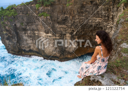 A beautiful woman in a pink dress sits on a cliff above the ocean on the island of Nusa Penida. Devil's Billabong an incredibly wonderful lagoon with splashes from the waves. 121122754