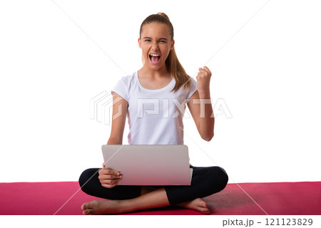 Emotional sporty young woman sitting in pink yoga mat in pose of lotus shout loud yeah fist up raise and using laptop , isolated on white background 121123829