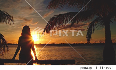 Silhouette of a young woman is sitting on a pier with palm trees at sunset. She is enjoying the view of a tropical lagoon Silhouette of a young woman is sitting on a pier with palm trees at sunset. She is enjoying the view of a tropical lagoon 121124951