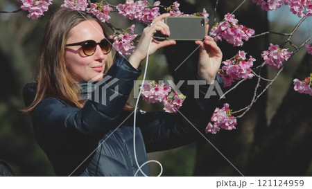 Young woman is taking pictures of beautiful cherry blossoms with her smartphone in a park in tokyo. She is enjoying the beautiful spring day 121124959
