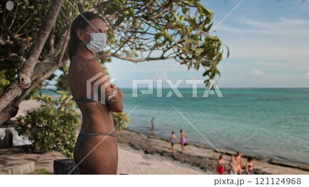 A lone woman wearing a face mask stands on a sandy beach, the vast ocean waves crashing behind her, symbolizing the surreal impact of COVID-19 on summer vacation. 121124968