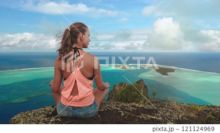 Young woman is practicing yoga and meditating, sitting on top of a mountain while admiring a scenic view of a tropical island 121124969