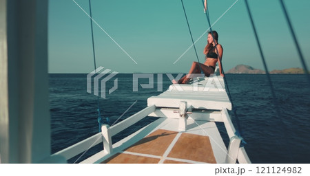 Woman relaxes on yacht sitting on wooden boat nose and looking at ocean wild nature landscape in background. Beautiful slim tanned girl in bikini has travel tour to Indonesia, Komodo National Park. 121124982