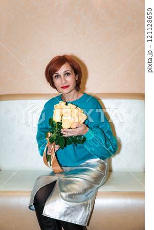 Russian woman, 48 years old, sitting indoors and holding a bouquet of yellow roses with a ribbon, wearing a turquoise blouse and silver skirt, posing with a calm and thoughtful expression Russian woman, 48 years old, sitting indoors and holding a bouquet of yellow roses with a ribbon, wearing a turquoise blouse and silver skirt, posing with a calm and thoughtful expression 121128650