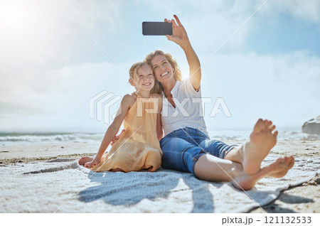 Carefree mother and daughter taking a selfie while sitting on the beach. Happy little girl and grandmother smiling while taking a picture on a cellphone while on holiday. Mom and daughter bonding 121132533