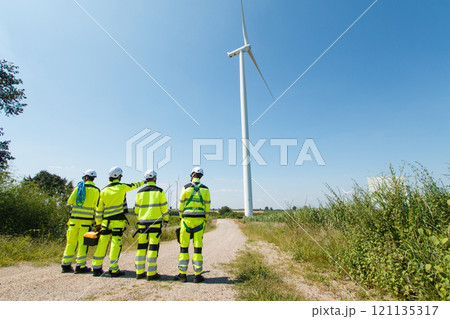 Team of professional electrical engineers in fully safety suit are working and discussing together at the windmill electric generating turbine. Electricians working at the site of windmill turbine. Team of professional electrical engineers in fully safety suit are working and discussing together at the windmill electric generating turbine. Electricians working at the site of windmill turbine. 121135317