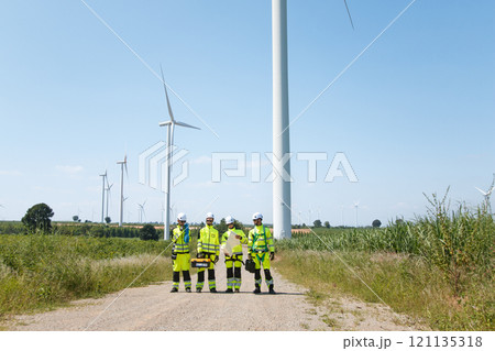 Team of professional electrical engineers in fully safety suit are working and discussing together at the windmill electric generating turbine. Electricians working at the site of windmill turbine. Team of professional electrical engineers in fully safety suit are working and discussing together at the windmill electric generating turbine. Electricians working at the site of windmill turbine. 121135318