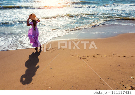 Woman in a straw hat with hands in the air walks on the beach. Young woman in a pink flattering dress on the seashore. Aerial view Woman in a straw hat with hands in the air walks on the beach. Young woman in a pink flattering dress on the seashore. Aerial view 121137432