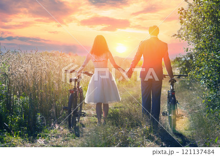 Young happy bride and groom with bicycles walk holding hands along the wheat field back to the camera during sunset Young happy bride and groom with bicycles walk holding hands along the wheat field back to the camera during sunset 121137484