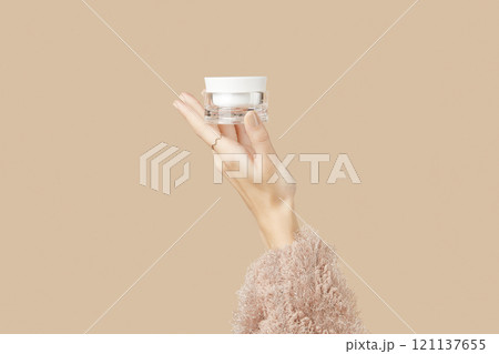 A womans hand in a sweater holds a jar of cream on a beige background. Body care beauty treatment A womans hand in a sweater holds a jar of cream on a beige background. Body care beauty treatment 121137655