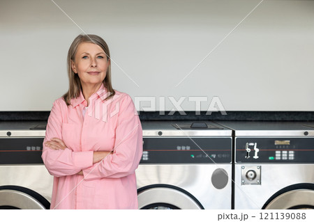 Confident mature woman in pink tshirt standing near washing machines in the laundry Confident mature woman in pink tshirt standing near washing machines in the laundry 121139088