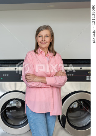 Confident mature woman in pink tshirt standing near washing machines in the laundry 121139090