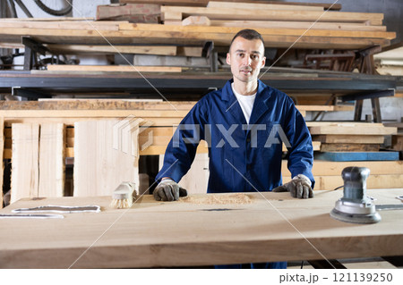 During manufacturing, man employee grinds surface of wooden board, gives smoothness to surface During manufacturing, man employee grinds surface of wooden board, gives smoothness to surface 121139250