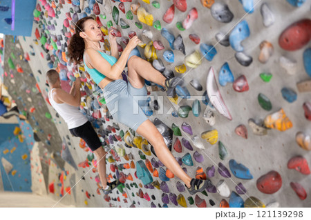 Woman in comfortable athletic clothing moving carefully on bouldering wall clinging to colorful hand and foot holds Woman in comfortable athletic clothing moving carefully on bouldering wall clinging to colorful hand and foot holds 121139298