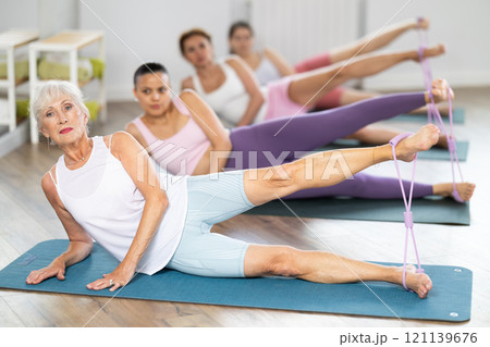 Elderly woman lies on mat and performs stretching exercises with resistance bands in group pilates class Elderly woman lies on mat and performs stretching exercises with resistance bands in group pilates class 121139676