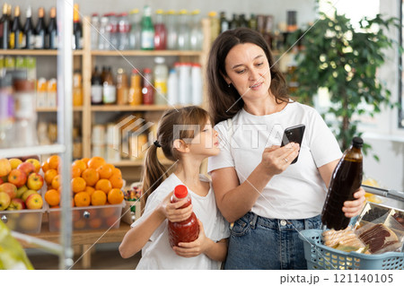 Mother and daughter choose bottles of juice and scan QR code with smartphone in supermarket 121140105