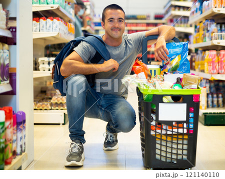 Cheerful man standing with full shopping basket in supermarket 121140110