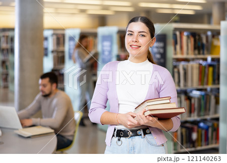 Smiling woman standing with books near bookshelves in library 121140263