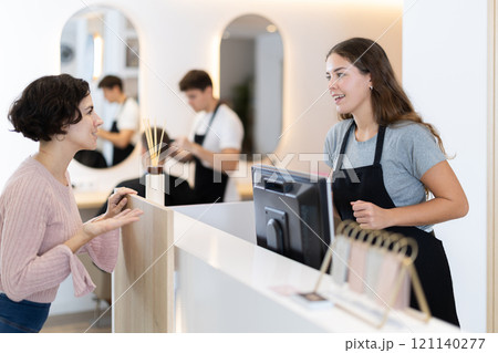 Female stylist stands near barbershop reception desk, communicates with female visitor 121140277