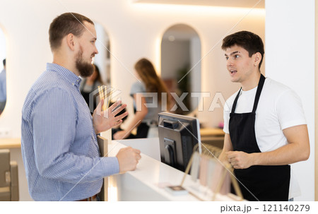 Male stylist stands near barbershop reception desk, communicates with male visitor 121140279
